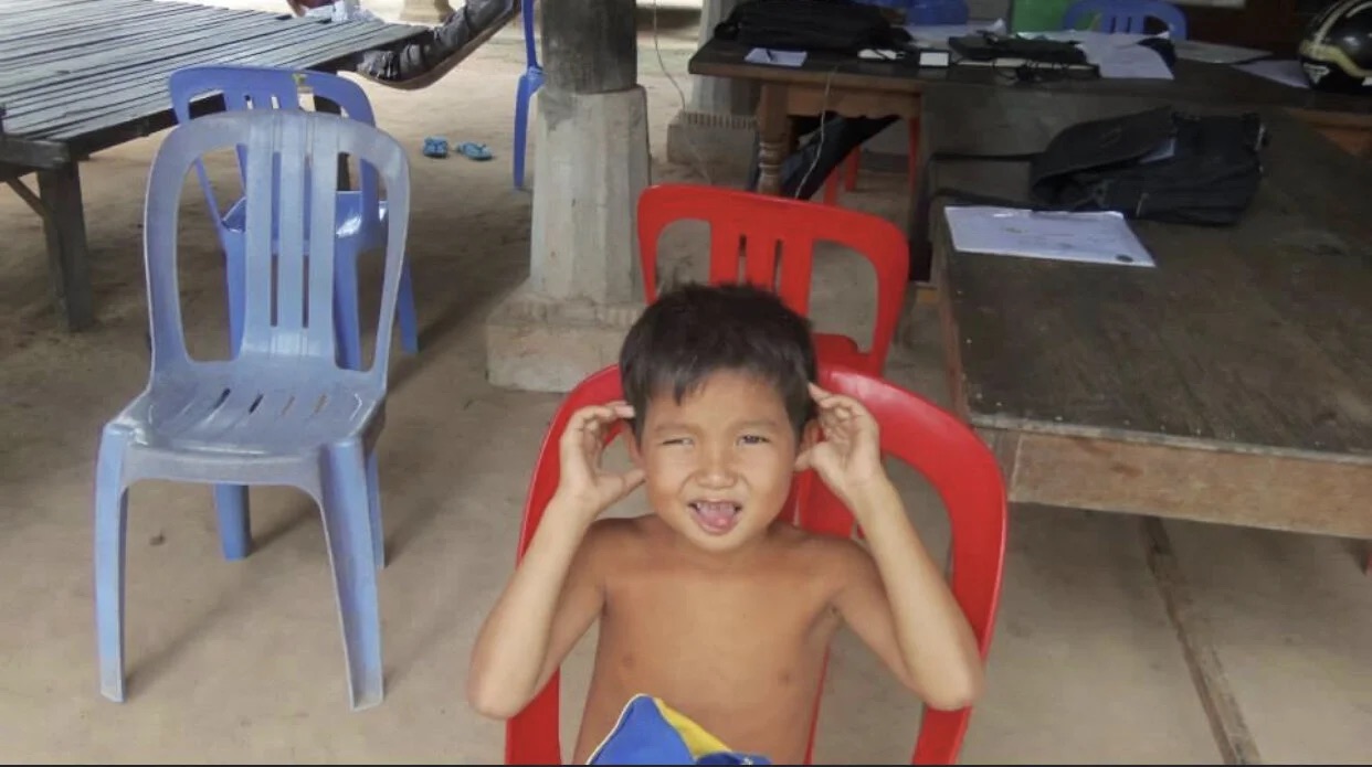 A young child sitting in a red plastic chair at a village workspace.